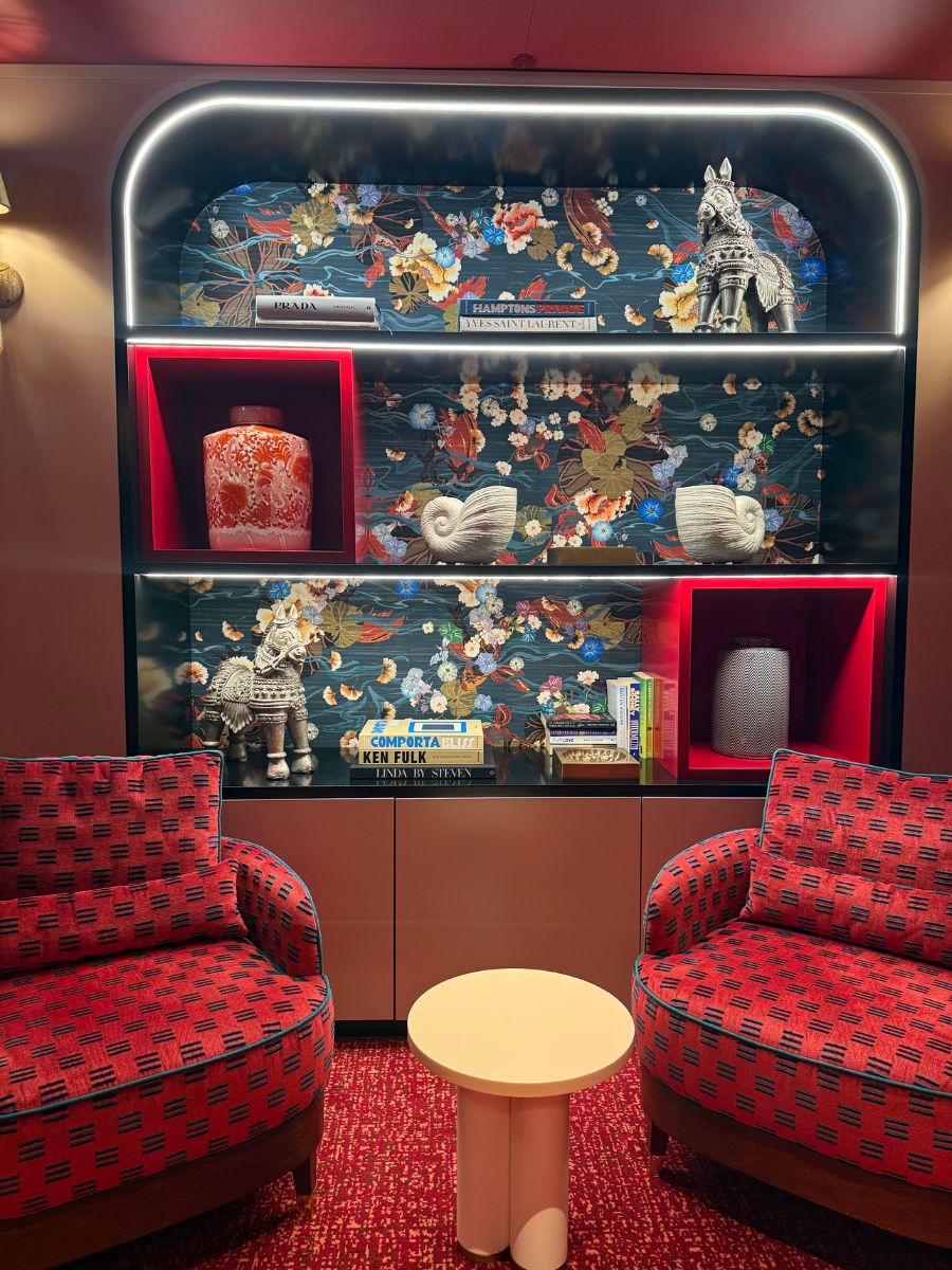 A cozy lounge area with two red-patterned armchairs, a round beige table, and a decorative wall shelf displaying books, vases, and statues, all set against a floral-patterned backdrop with accent lighting.