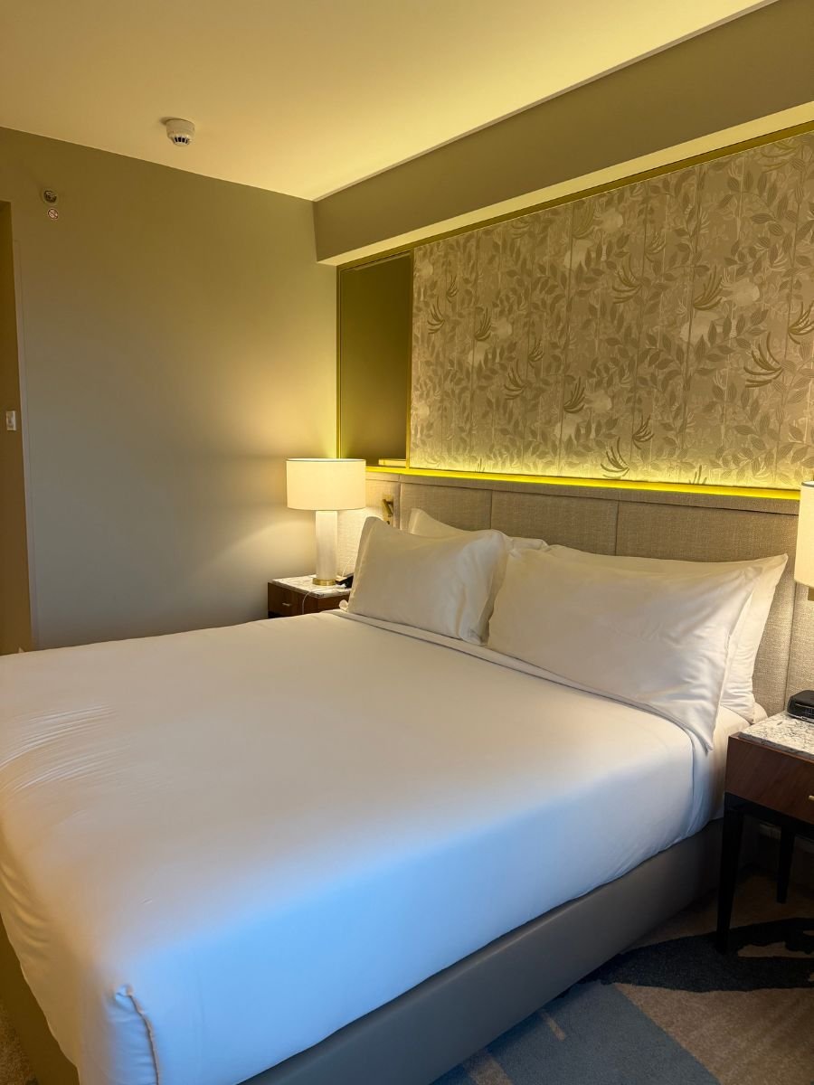 A modern hotel room with a neatly made king-sized bed, two bedside tables with lamps, and a softly lit, textured wall panel behind the headboard.