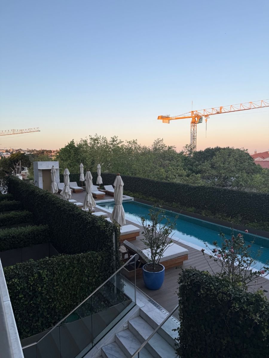 An outdoor pool area with white lounge chairs and umbrellas, surrounded by manicured hedges and trees, with construction cranes visible in the background under a clear sky.