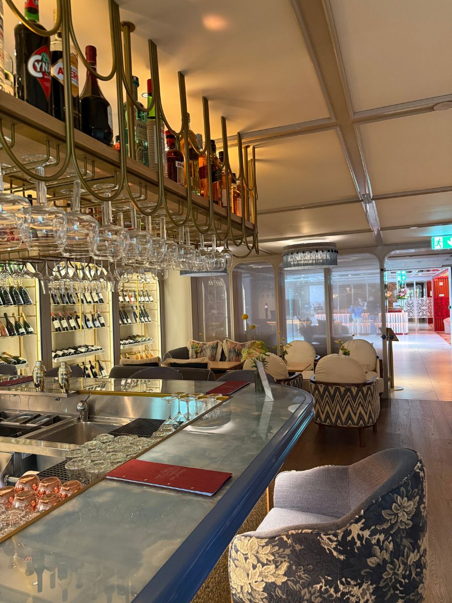 A stylish hotel bar with a hanging glass rack, bottles of liquor, and plush armchairs, with natural light coming through large windows and a wine display on the back wall.
