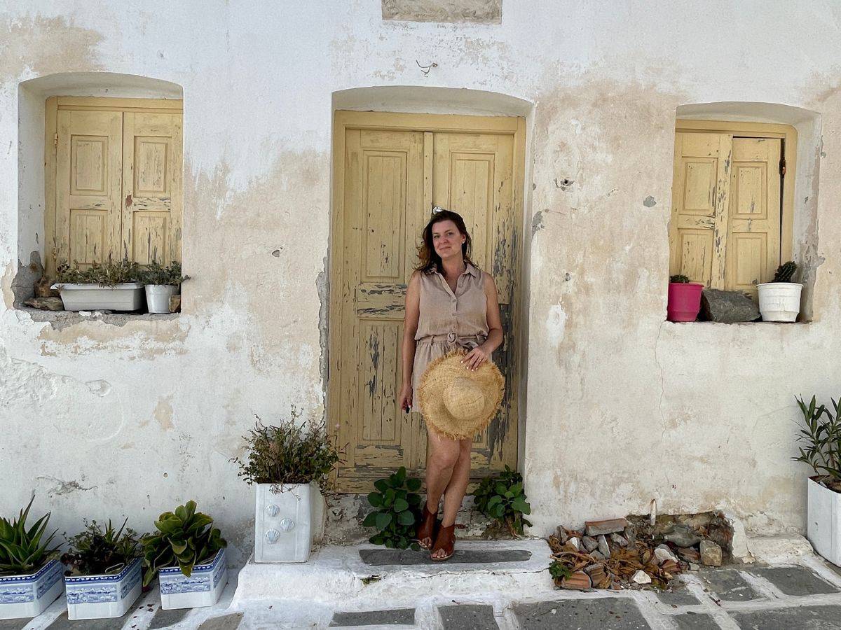 A solo female traveler stands confidently in front of a rustic, white-washed wall with weathered wooden doors in a charming old town. She is dressed in a casual, chic sleeveless dress, paired with comfortable sandals, perfect for exploring. She holds a straw hat, a symbol of her readiness for adventure. Potted plants add a touch of greenery to the scene, enhancing the vibe of a tranquil getaway. This image embodies the spirit of overcoming the fear of traveling alone, showcasing the joy of solo adventures and the beauty of quaint, undiscovered spots