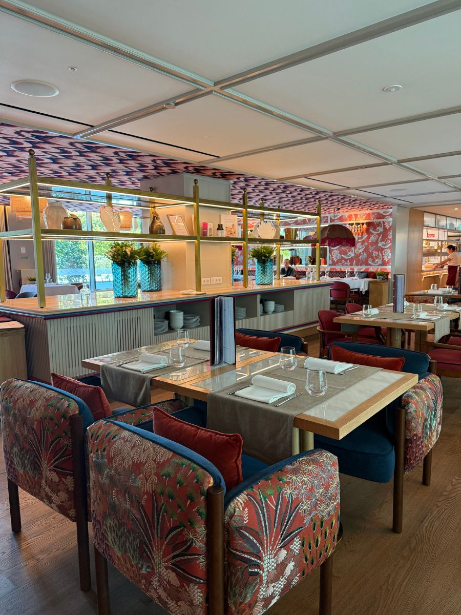 A vibrant hotel restaurant with colorful upholstered chairs, neatly set tables with glassware and napkins, and decorative shelving with vases and plants separating dining spaces.