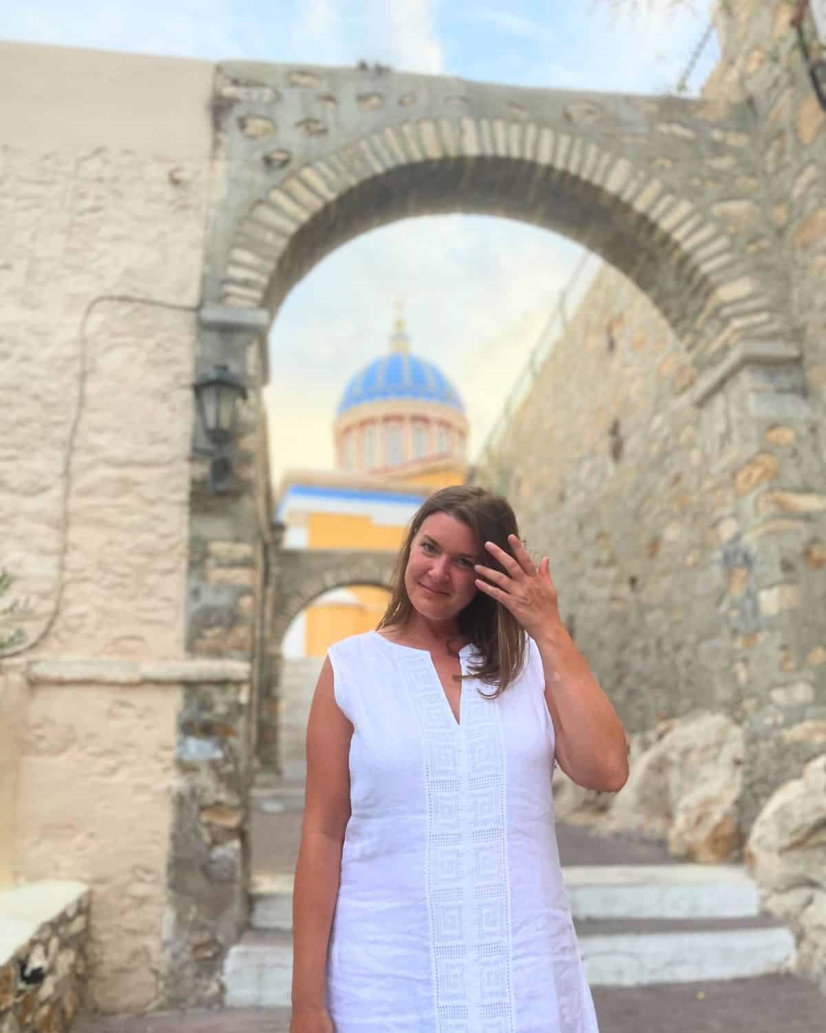 A woman wearing a blue and white patterned dress stands confidently with her hands resting behind her head on a rocky shoreline, with the calm sea behind her and a coastal town in the distance. The clear sky and her relaxed posture suggest a sense of freedom and joy in solo travel.