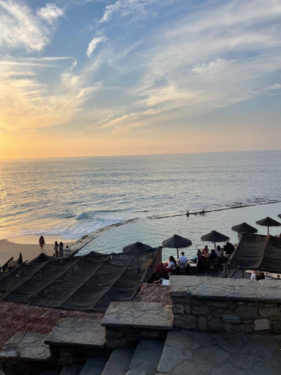 Stunning sunset view at Azenhas do Mar, with guests enjoying seaside dining, umbrellas silhouetted against the golden glow of the horizon.