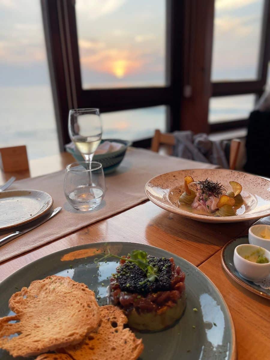 Dining table with seafood dishes and a glass of wine, set against the backdrop of a soft, ocean sunset seen through the windows.