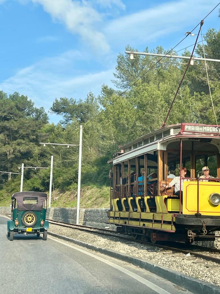 Charming historic yellow tram traveling towards Praia das Maçãs alongside a vintage car, framed by lush green trees.