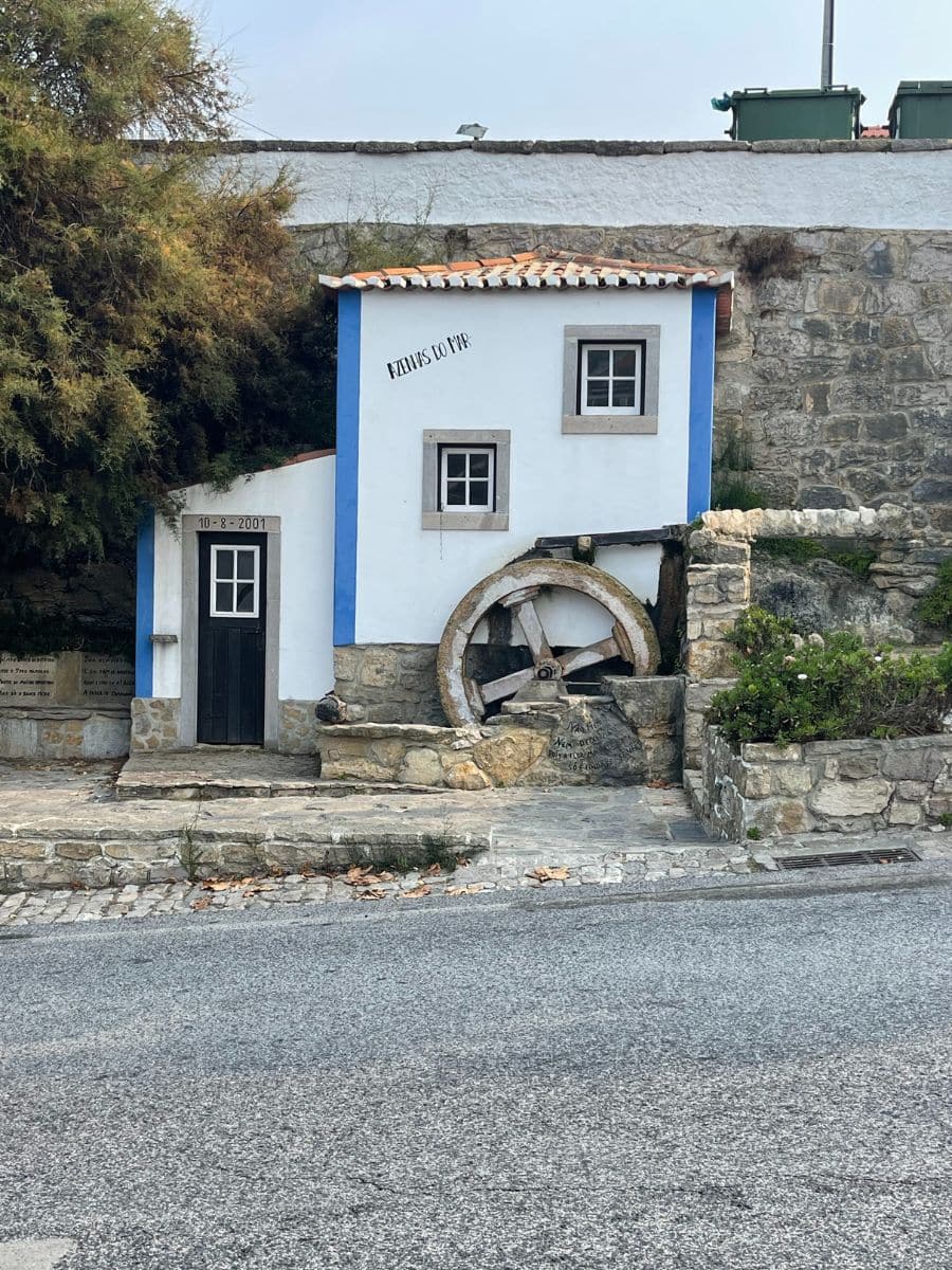 Charming white-and-blue historic water mill building, known as 'Azenhas do Mar', featuring a rustic wooden water wheel.