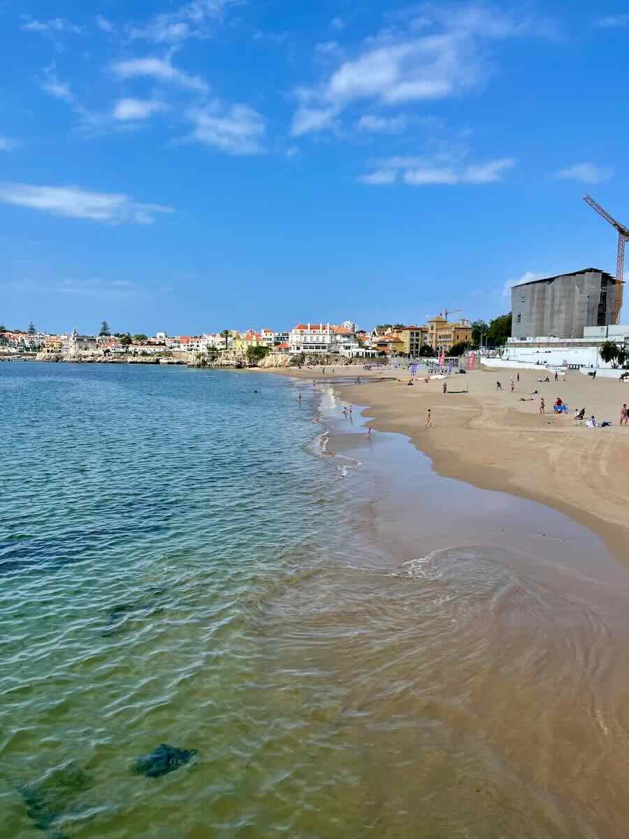 A sandy beach in Cascais, Portugal, with calm blue-green waters gently touching the shoreline. In the background, picturesque seaside buildings and a few visitors enjoying the beach under a bright blue sky.