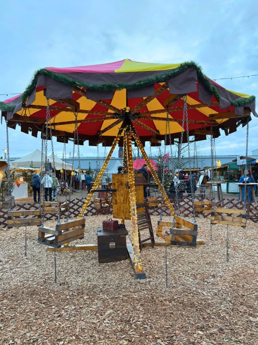 A colorful carousel with wooden swing seats decorated in bright red and yellow, surrounded by wood-chip ground at a Christmas market at RAW Gerlide Berlin.