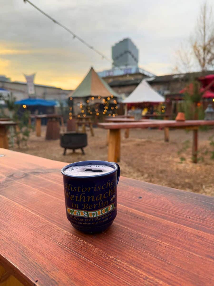 A blue souvenir mug of glühwein resting on a wooden table, with festive market tents and twinkling lights in the background during sunset.