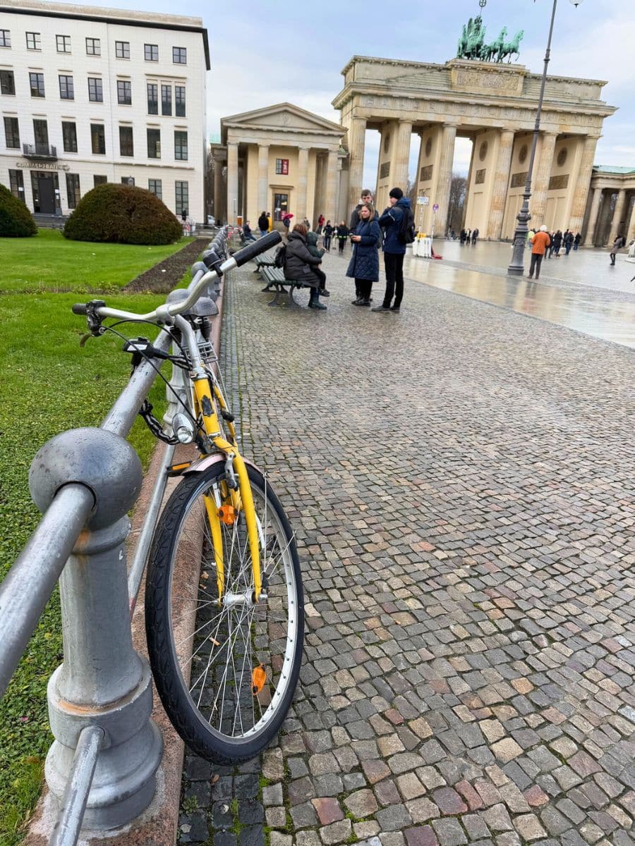 A bright yellow bicycle leaning against a railing in front of the iconic Brandenburg Gate, surrounded by tourists and cobblestone streets in Berlin.