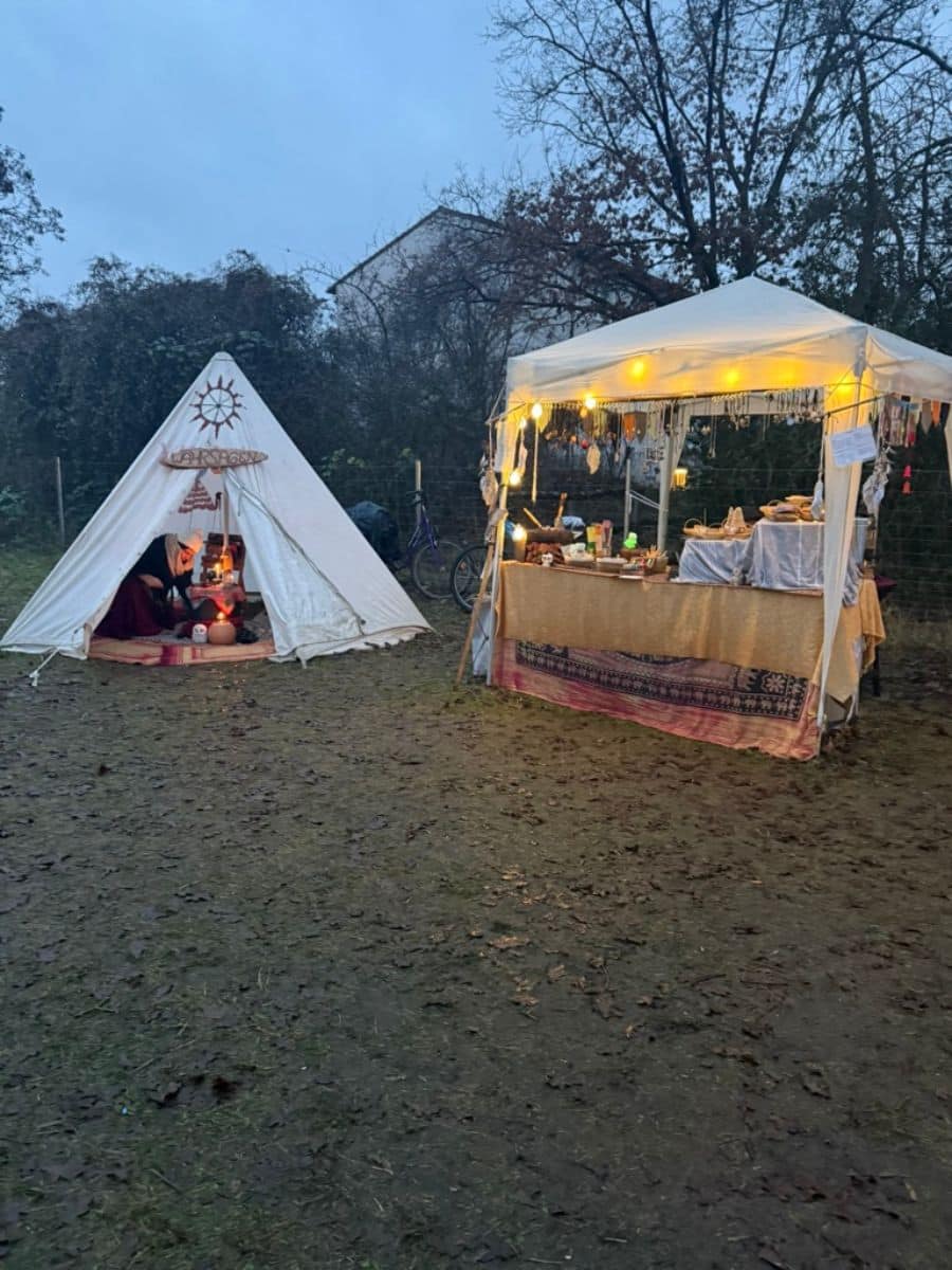 A cozy scene with a small triangular tent decorated with festive items and a brightly lit craft stall, set on the grassy grounds of Britz Castle.