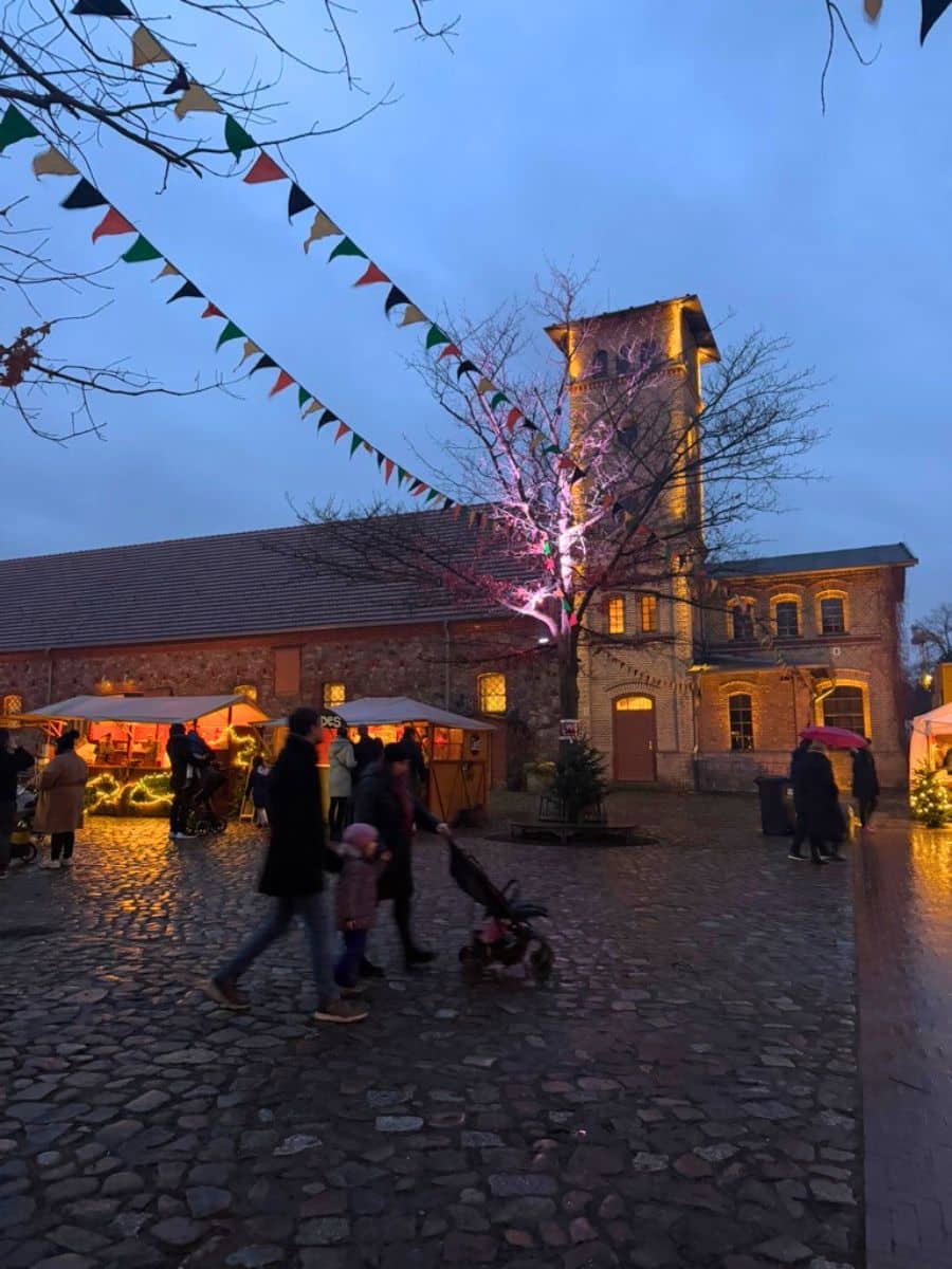The illuminated Britz Castle tower and cobblestone courtyard surrounded by market stalls, with visitors strolling under strings of colorful bunting.