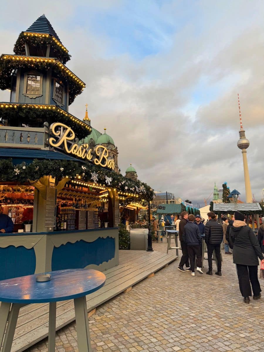 A festive Christmas bar decorated with garlands and lights at the Humboldt Forum Christmas Market, with the Berlin TV Tower visible in the background.