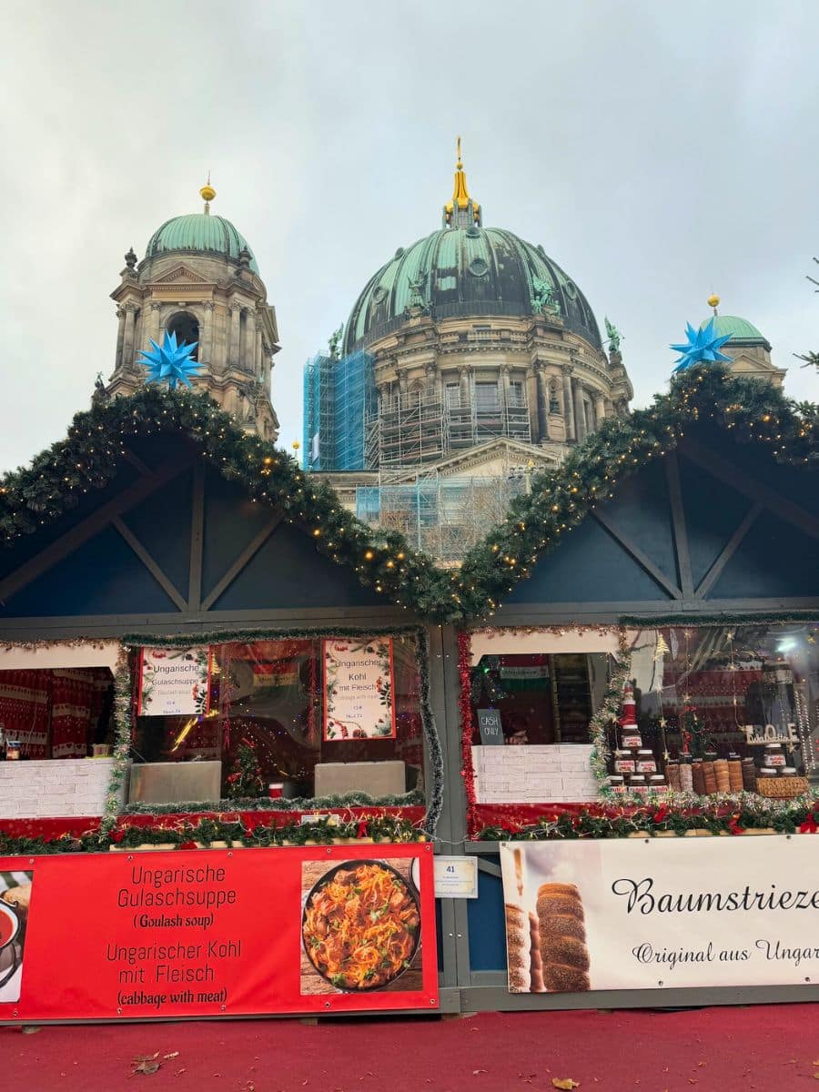 A food stall decorated with Christmas greenery, offering Hungarian specialties like goulash soup and chimney cakes, with the Berlin Cathedral domes towering above.