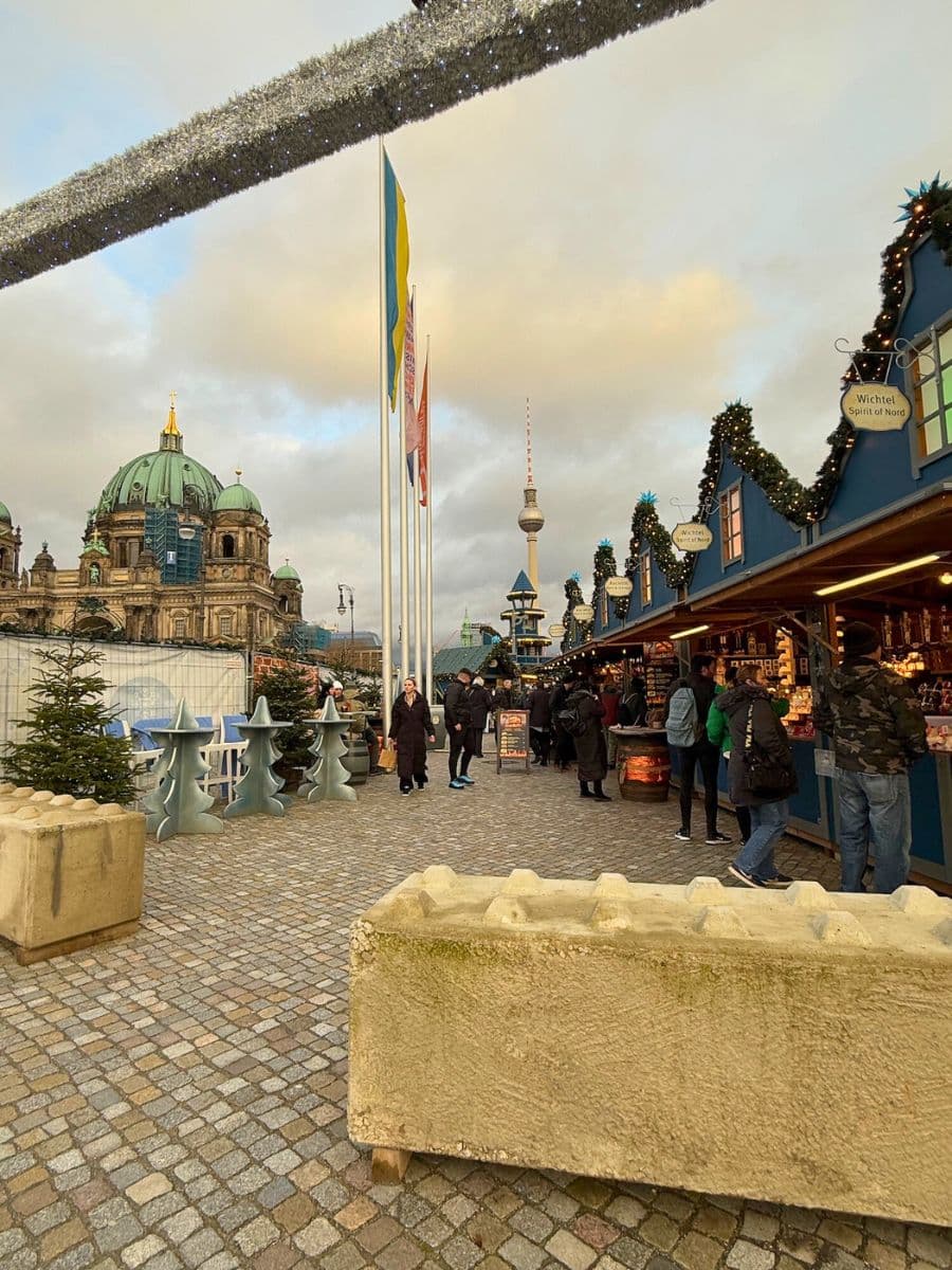 A wide view of the Humboldt Forum Christmas Market with festive stalls, decorative garlands, and the Berlin Cathedral and TV Tower in the background. Large concrete barriers are visible at the market's entrance for safety.