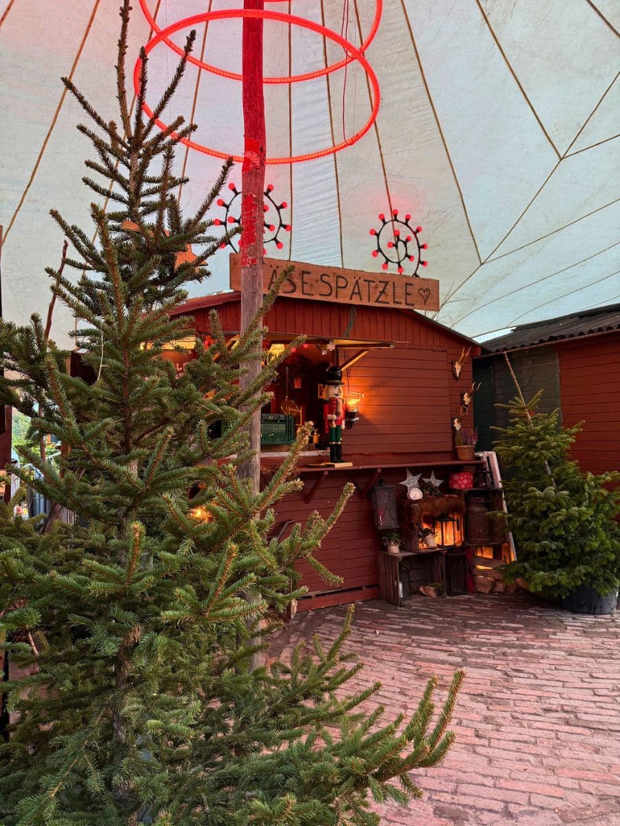 A cozy market stall named "Käse Spätzle" under a large canopy, decorated with festive lights and Christmas trees, offering warm winter food.