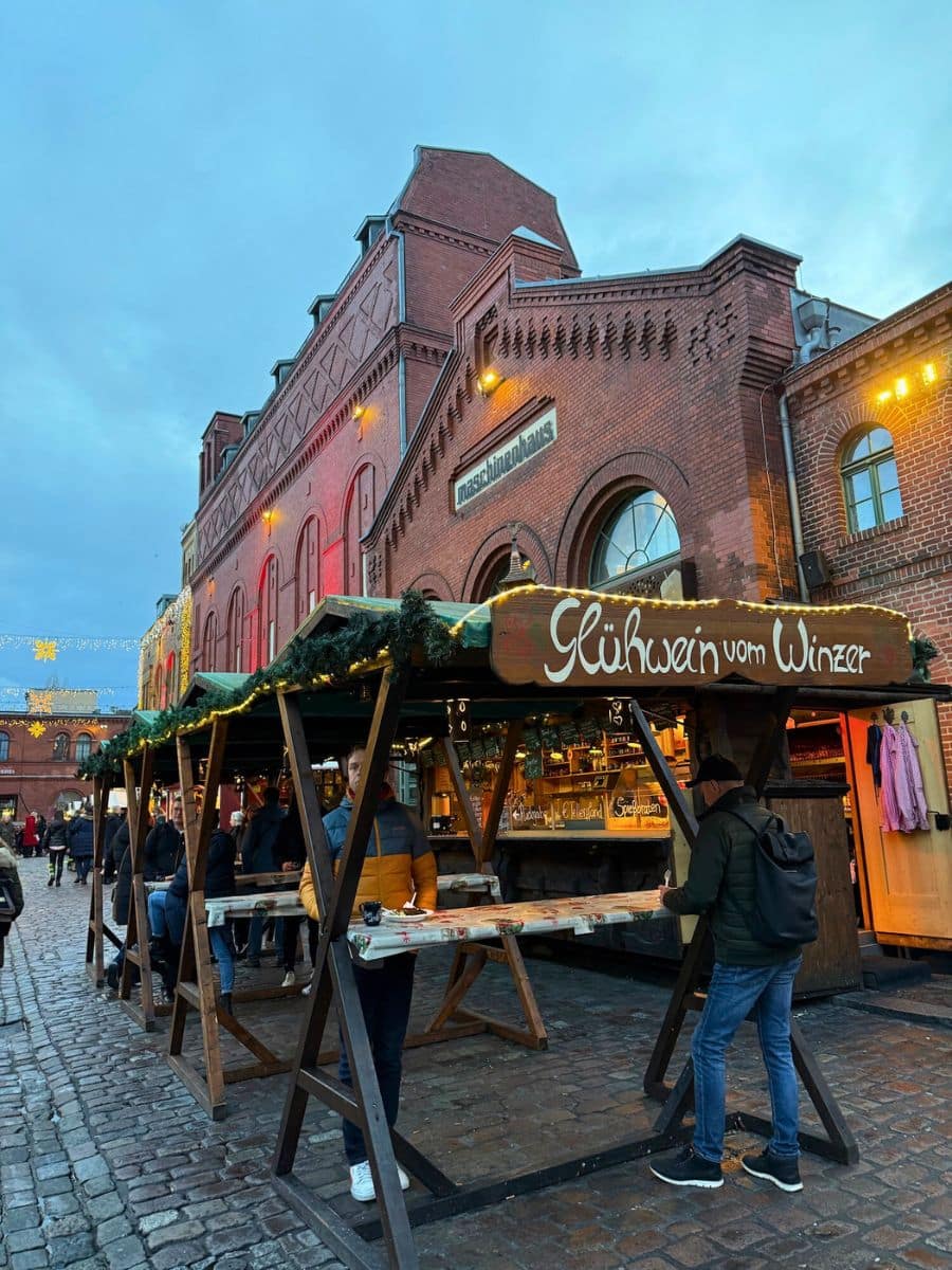 A traditional glühwein stall with rustic wooden seating and the iconic brick facade of the Berlin Markthalle in the background, lit with warm festive lights.
