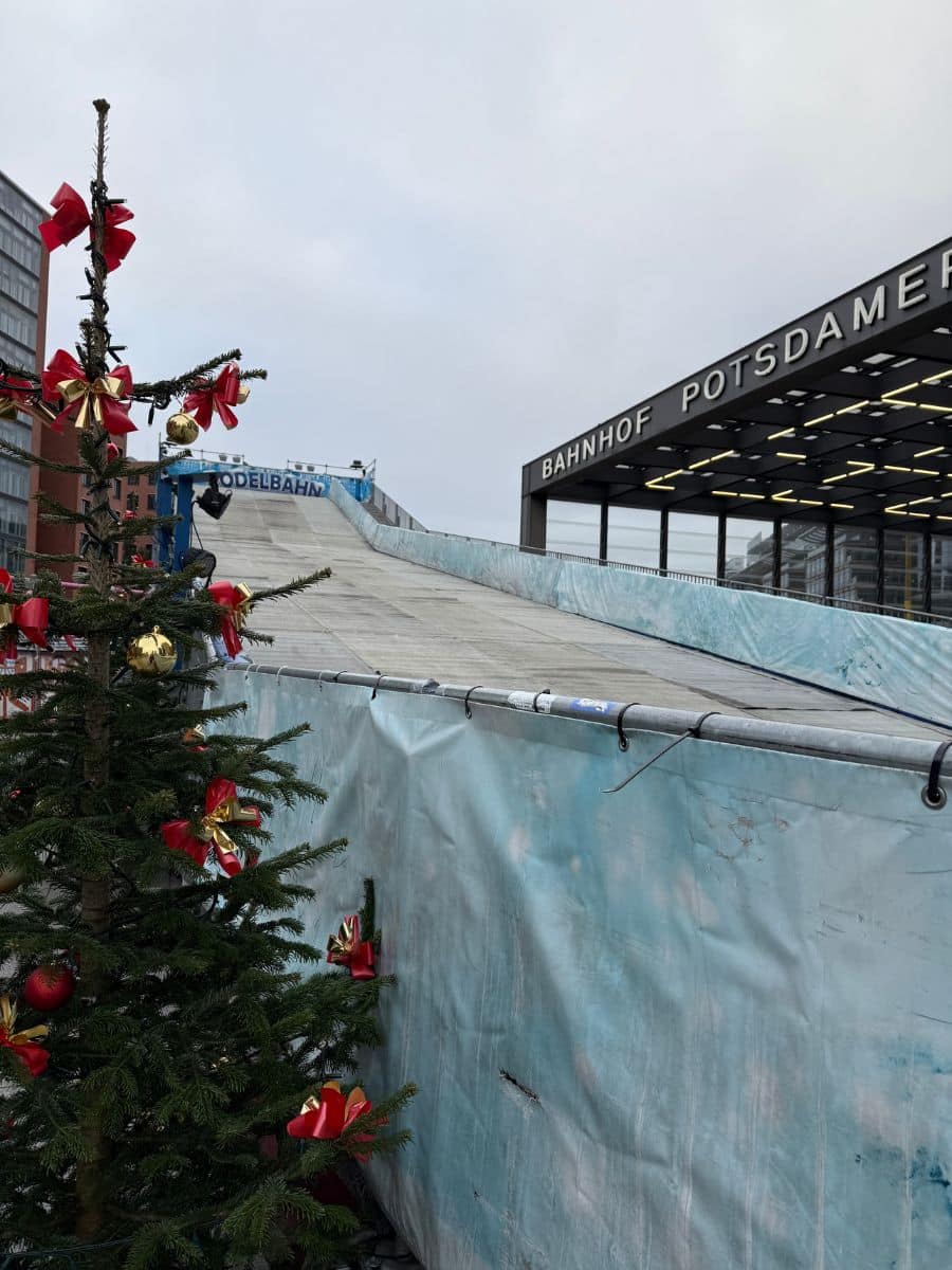 A decorated Christmas tree framing the view of a sledding hill at Potsdamer Platz, Berlin, with the sign "Bahnhof Potsdamer Platz" visible in the background.