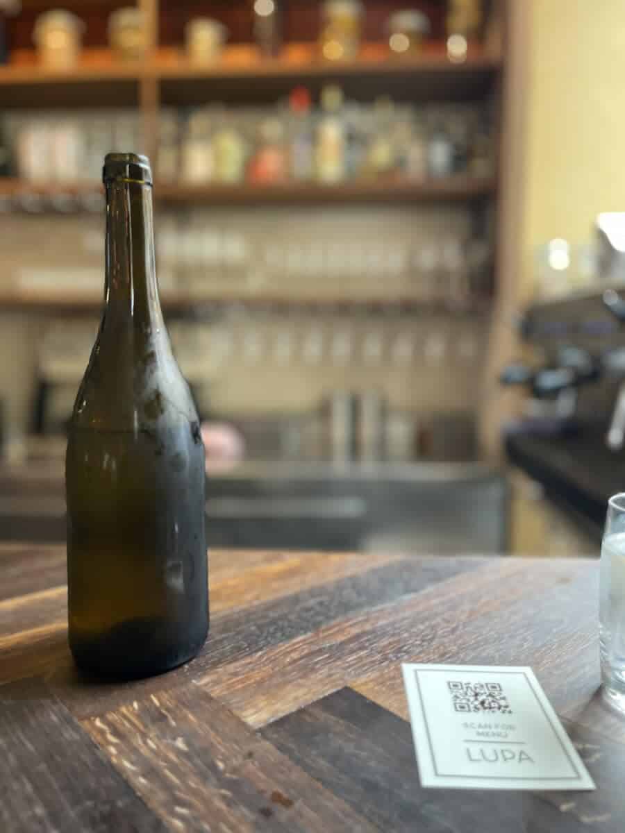 A close-up view of a wine bottle on a wooden table in a cozy Copenhagen bistro, with a blurred background of shelves stocked with bottles.