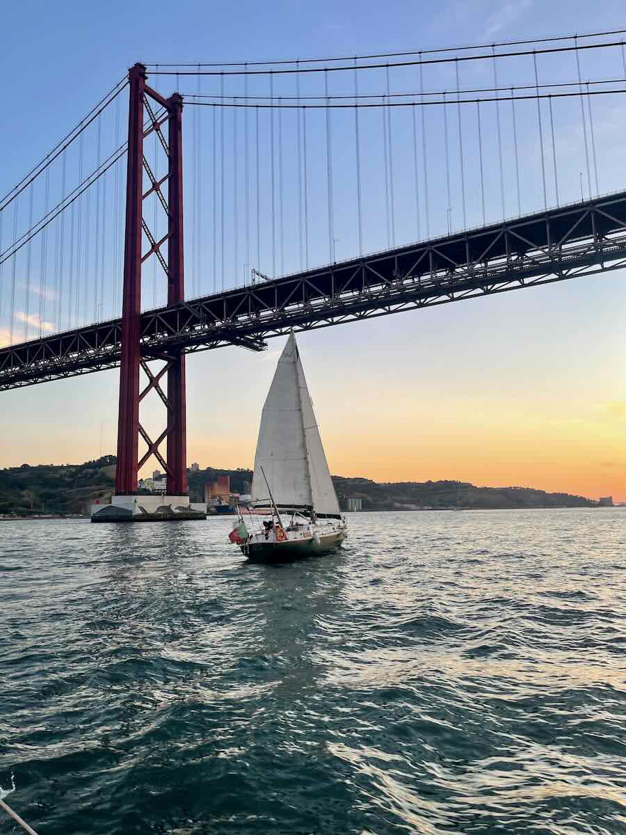A sailboat with a tall mast glides under a large red suspension bridge at sunset. The water ripples gently, and the sky transitions from blue to a soft orange glow, casting reflections on the water. The bridge’s structure looms above, framing the serene scene.