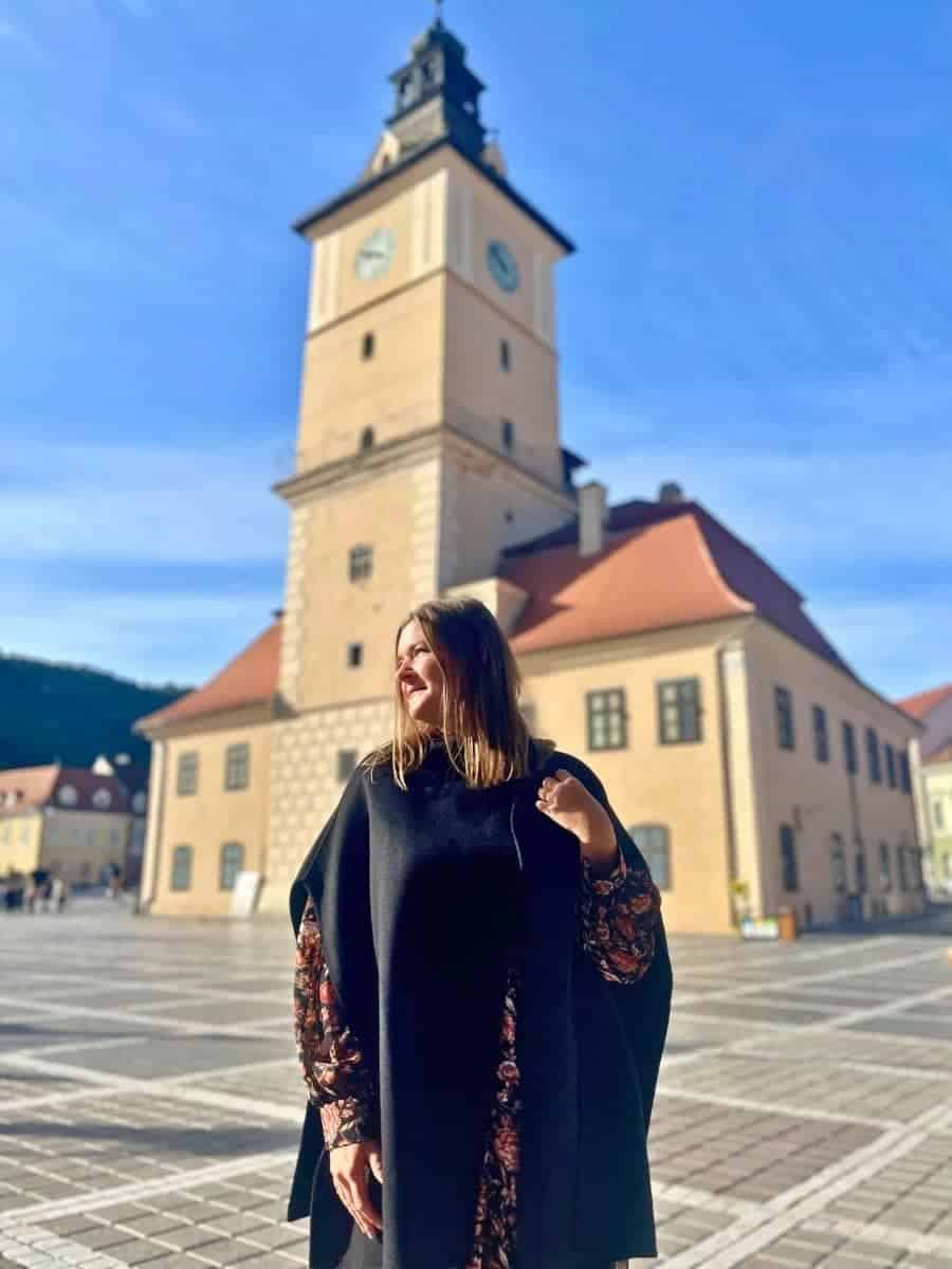 Solo female traveler standing in Bra?ov's main square, Piata Sfatului, with the iconic Council House and clock tower in the background on a bright, sunny day.