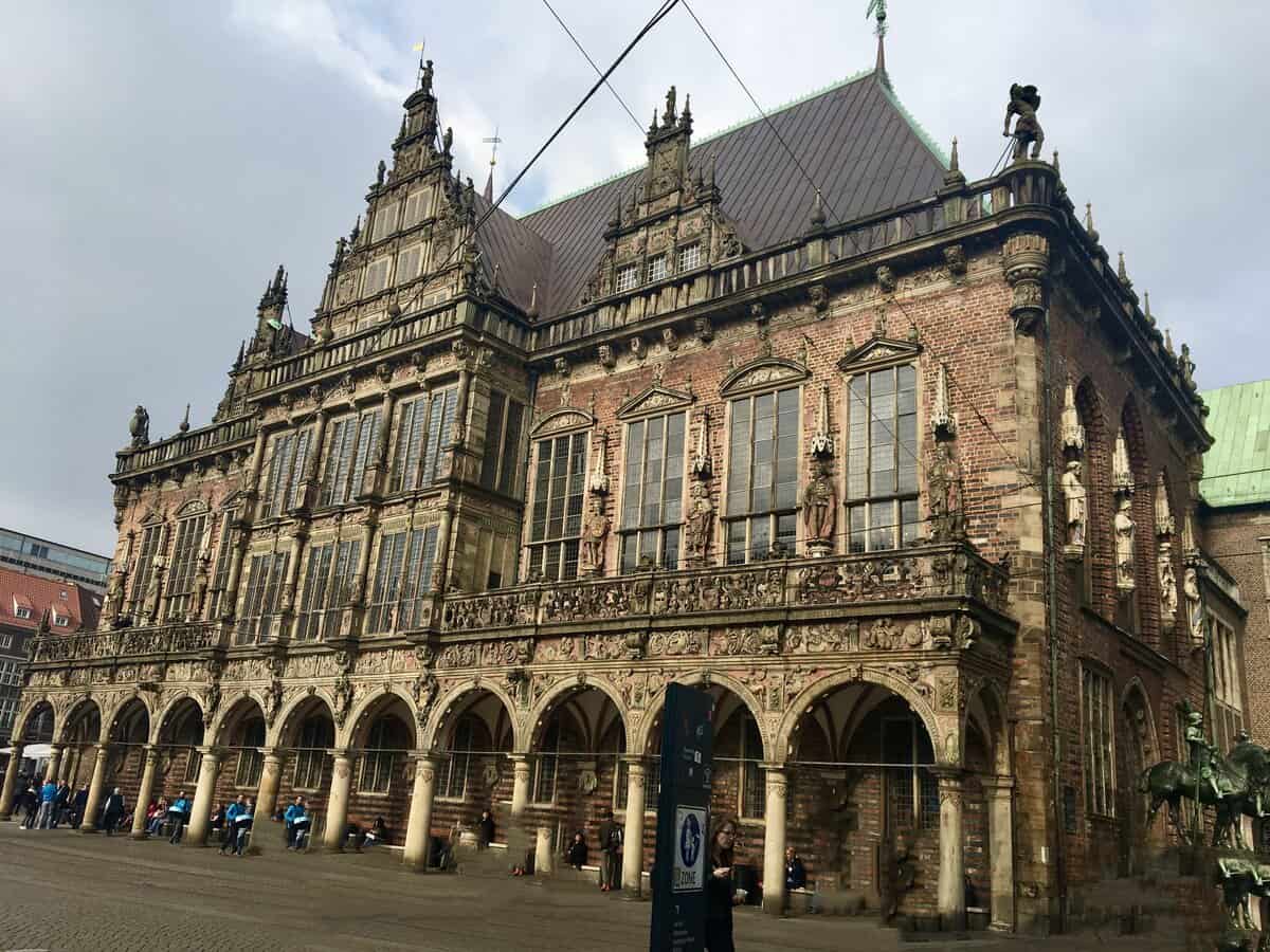 The Bremen Town Hall, a Renaissance-style building with intricate façade decorations including statues and reliefs. The exterior combines sculpted stone and brickwork, with large windows and a series of arches at ground level providing shelter to people below. 