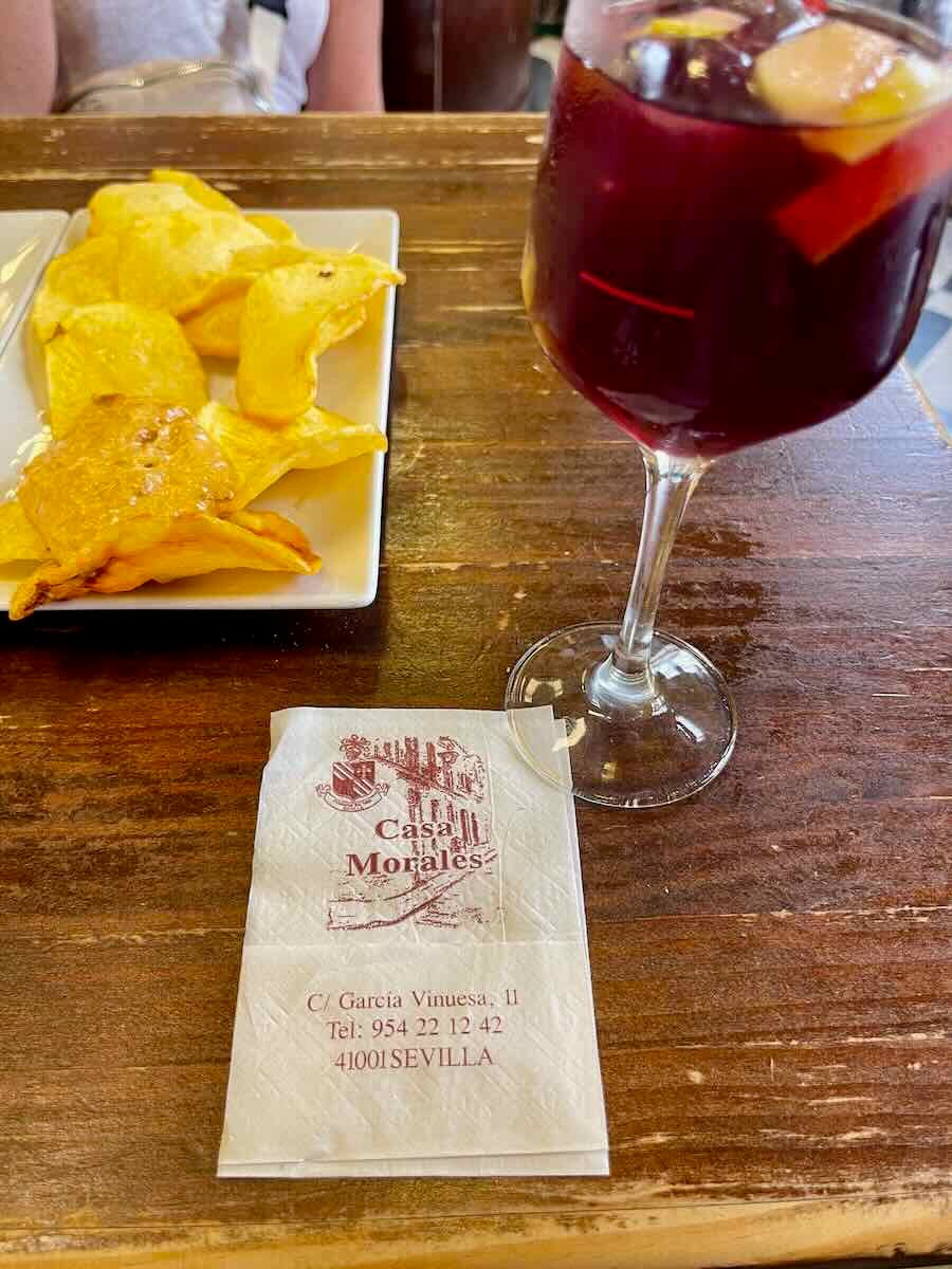 A close-up photo of a table at Casa Morales, a bar in Seville, Spain. The image shows a glass of sangria with fruit pieces floating in it, a small plate of potato chips, and a napkin with the bar's logo and address printed on it.