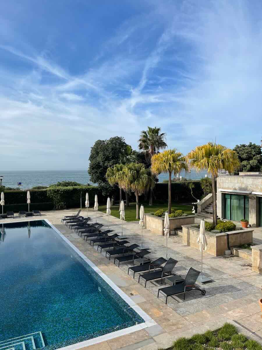 Serene outdoor hotel pool area with rows of lounge chairs and umbrellas, surrounded by lush greenery and palm trees, overlooking the ocean on a clear, sunny day.