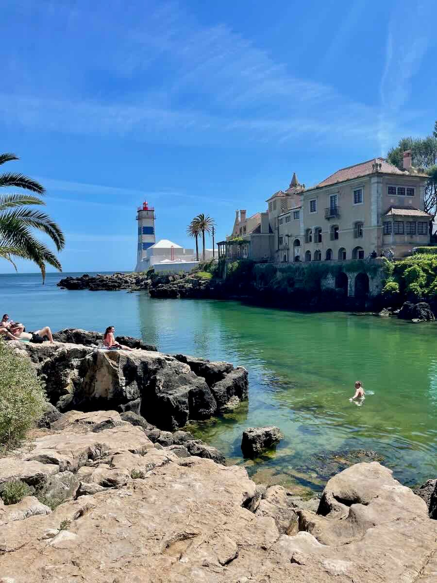 A beautiful scene in Cascais, Portugal, featuring a cove with clear green waters, rocky outcrops, and people relaxing by the sea. A striped lighthouse stands in the background, framed by palm trees and a historic building, all set under a clear blue sky.
