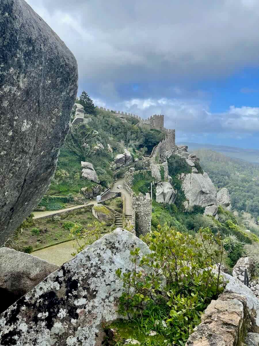 A dramatic view of the ancient castle walls in Sintra, Portugal, winding across a green hilltop. Large boulders frame the scene, and the lush landscape stretches into the distance under a partly cloudy sky.