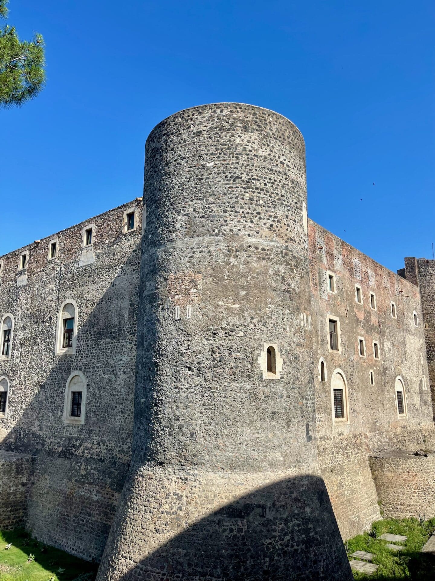 Exterior view of the Norman Castello Ursino in Catania, Italy, featuring its impressive medieval stone walls and circular tower under a bright blue sky.