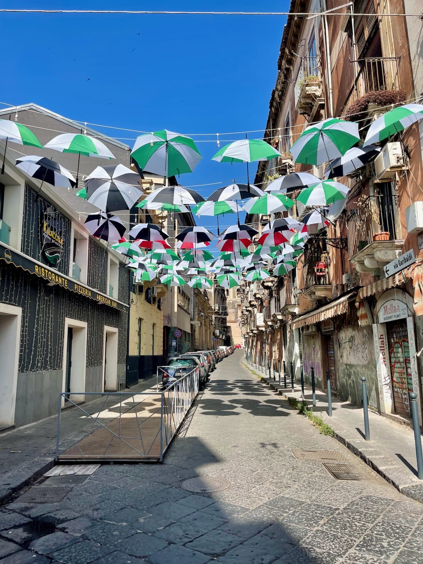 A street in Catania decorated with hanging umbrellas in various colors, under a clear blue sky.
