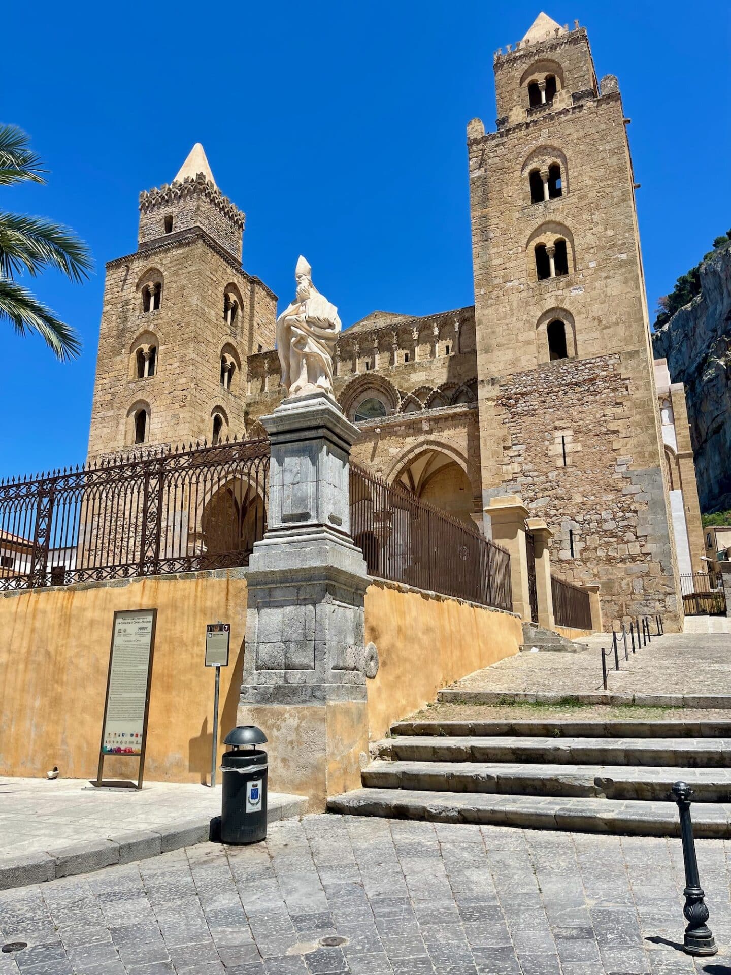 Cefalù Cathedral with two tall square towers and a statue of a bishop in front, under a clear blue sky.