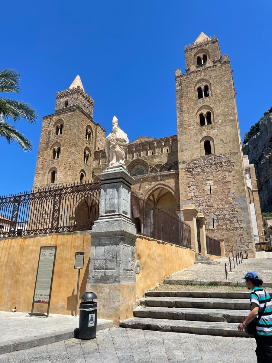 The Cathedral of Cefalù, a majestic Norman-style church in Sicily, stands against a vibrant blue sky. The structure features two imposing bell towers with arched openings and a grand entrance with intricate arches. A white marble statue of a religious figure is positioned on a stone pedestal in front of the cathedral. A set of stairs leads up to the main entrance, with an iron fence enclosing the area. To the left, an informational sign provides historical details about the site. A palm tree partially frames the left side of the image, while a pedestrian wearing a striped shirt and a cap walks in the foreground. The clear blue sky enhances the beauty of this UNESCO-listed landmark.