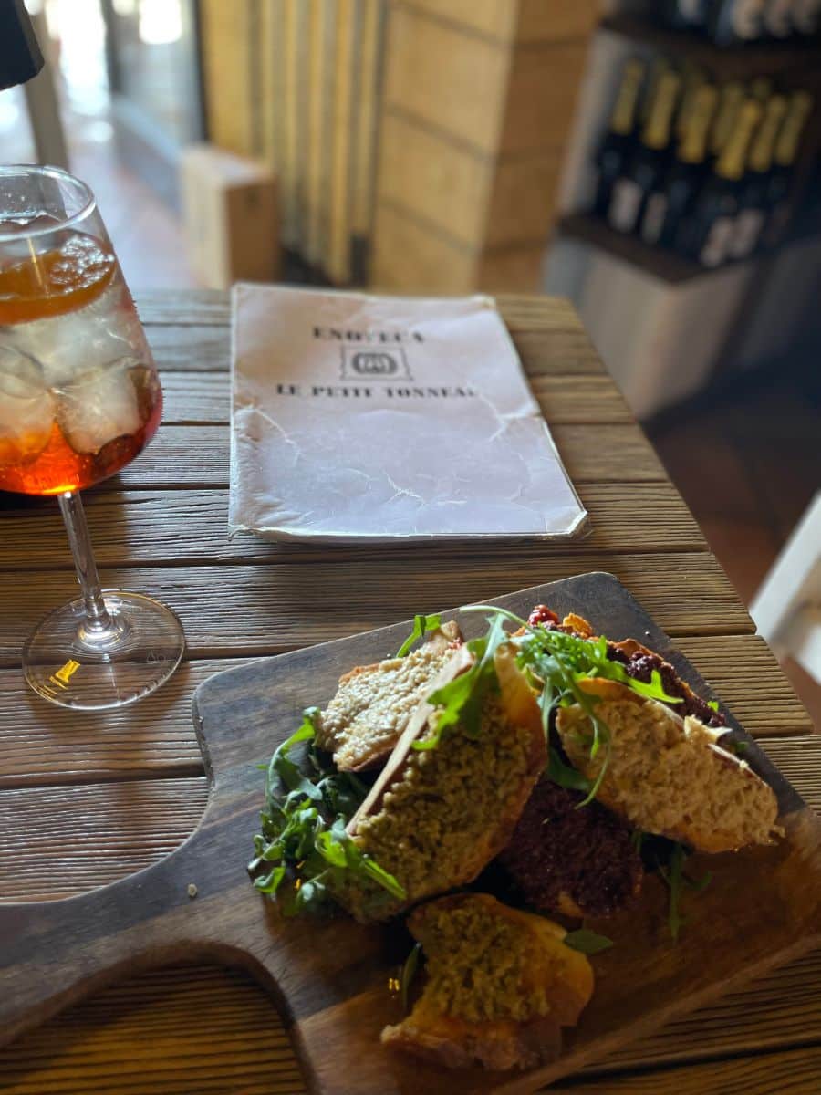 A close-up of a wooden table in a cozy wine bar, featuring a rustic cutting board with slices of bread topped with various spreads, garnished with fresh arugula. A chilled glass of Aperol Spritz with ice sits beside the dish. In the background, a slightly crumpled menu labeled 'Enoteca Le Petit Tonneau' rests on the table, with a shelf filled with wine bottles visible further back. The setting exudes a warm and intimate atmosphere, perfect for enjoying Sicilian flavors.