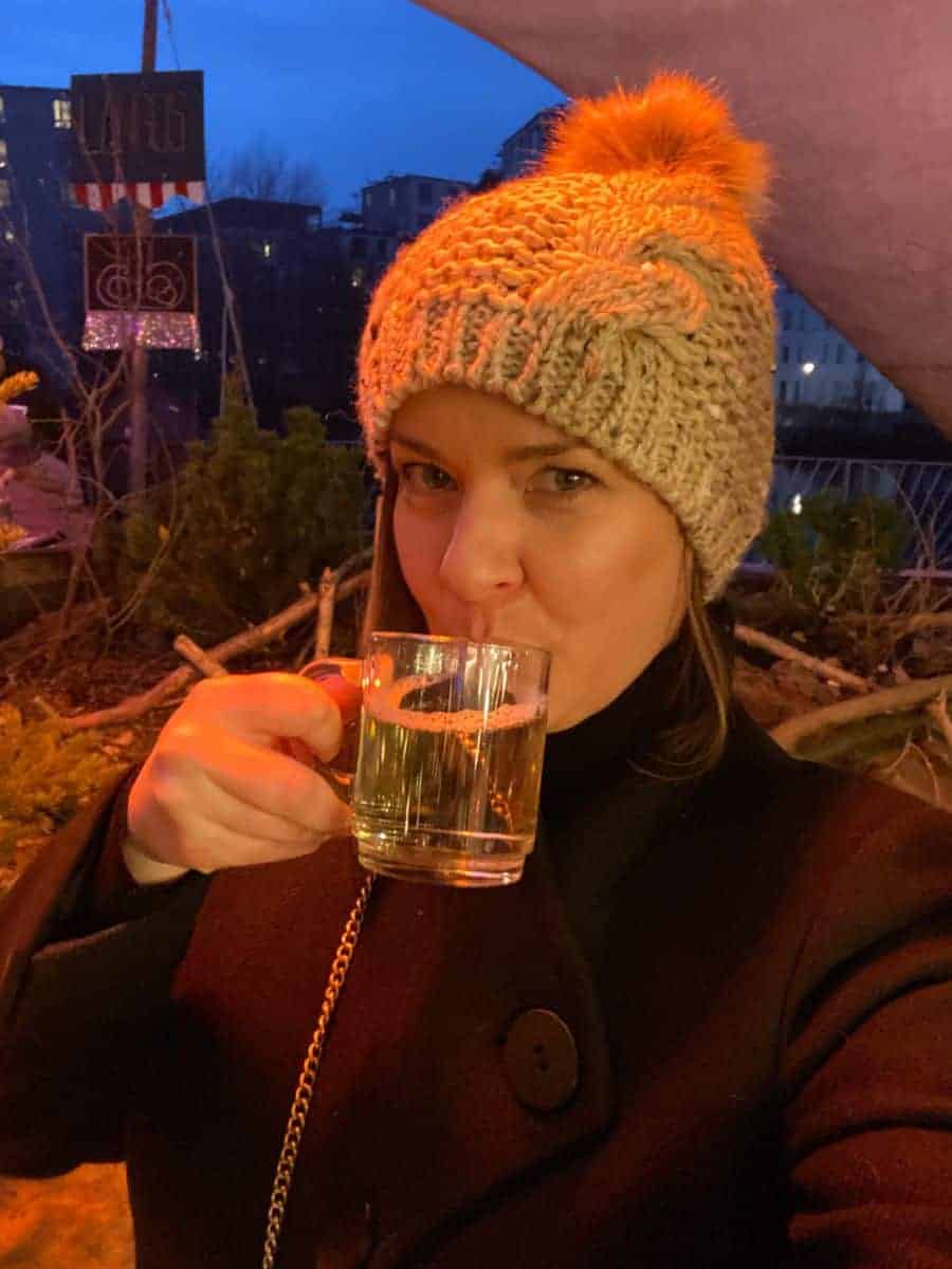 A woman in a warm knit hat with a pom-pom and a black coat is sipping from a clear glass mug, likely enjoying a warm drink at an outdoor setting in the evening.