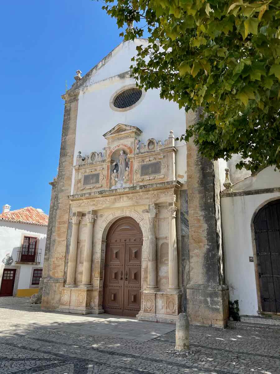 A historic church in Óbidos, Portugal, featuring a beautiful stone facade with intricate details around the doorway, columns, and statues. A large wooden door stands in the center, while the building is framed by lush green tree branches and a clear blue sky.