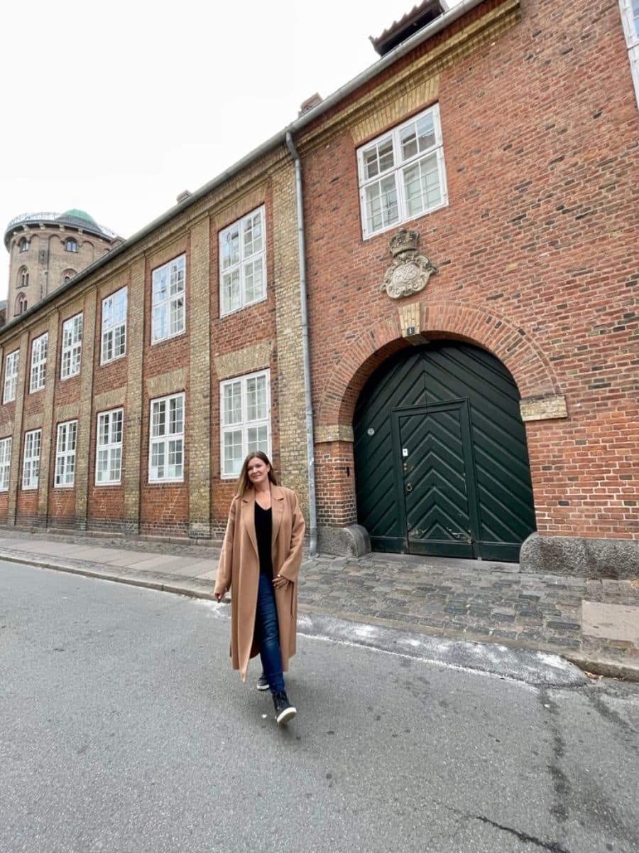 A woman in a long beige coat strolling past an historic brick building with a large wooden door in Copenhagen, illustrating the blend of old architecture and modern life in the city.