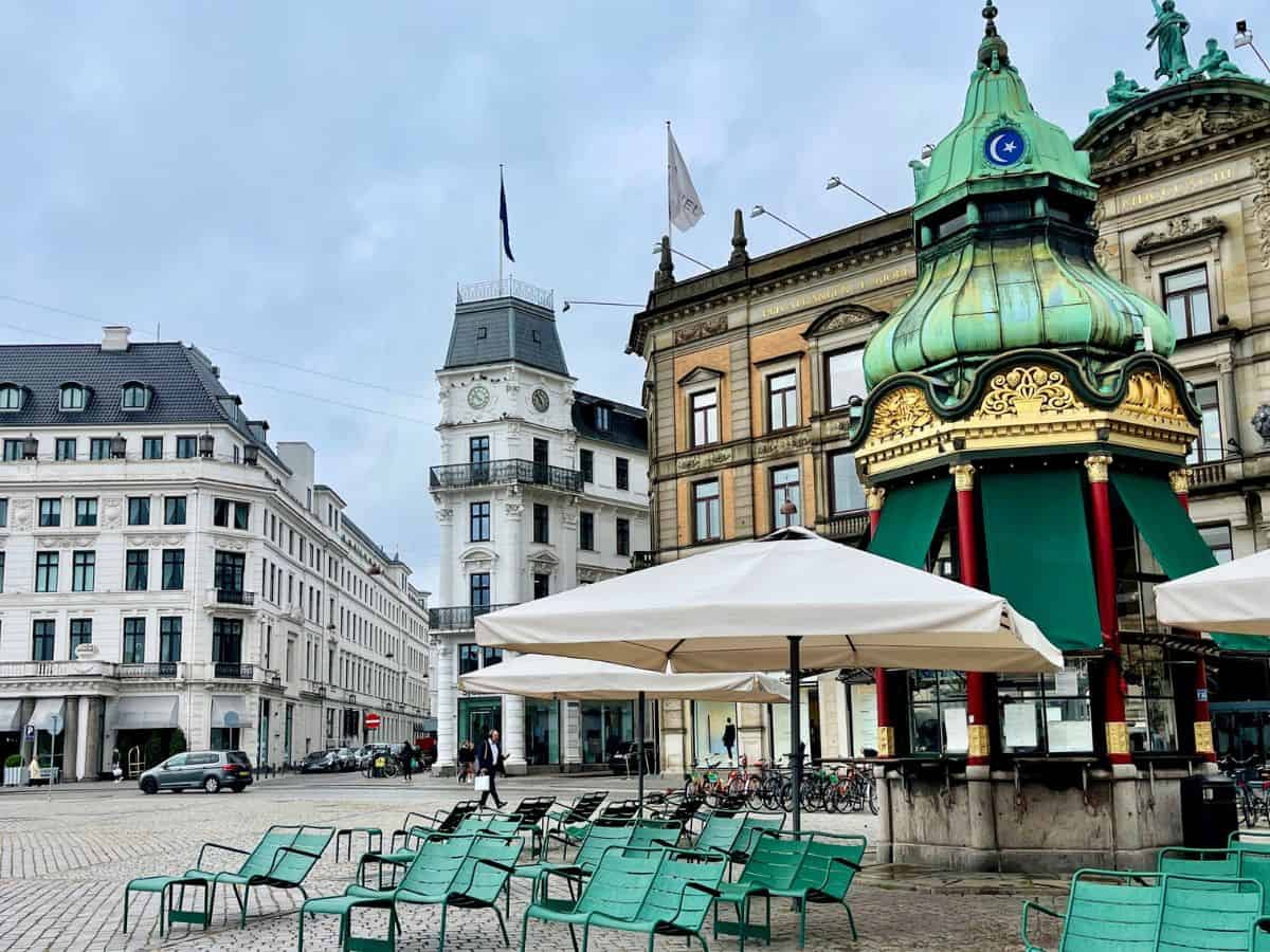 The image shows a quiet square in Copenhagen with a green-topped pavilion, surrounded by classic European buildings. Green chairs and tables are set up under umbrellas, suggesting a peaceful outdoor café scene.
