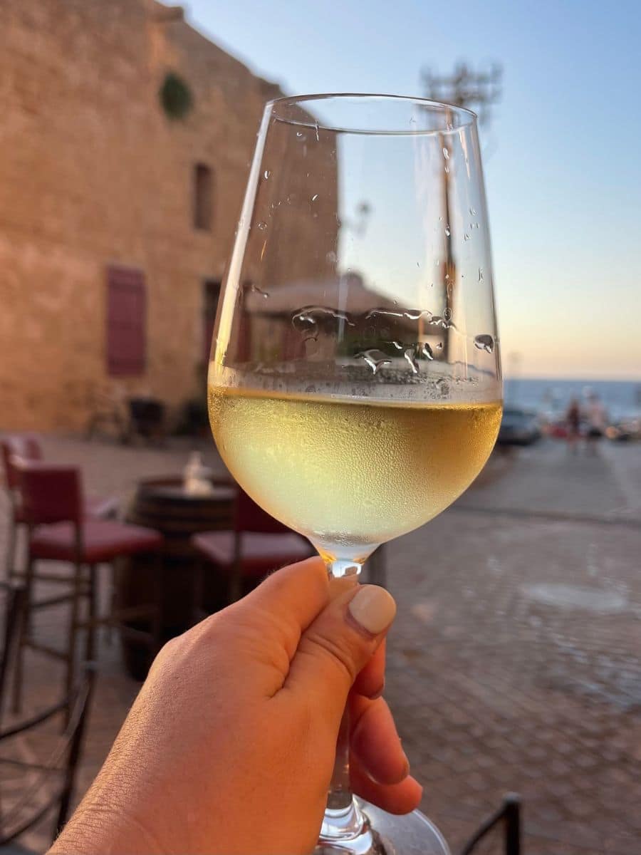 Close-up of a chilled glass of white wine held in a hand, with a blurry background of a historic European street at sunset, epitomizing leisurely travel moments.
