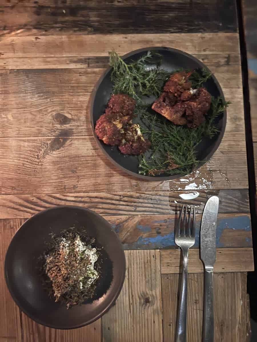 A rustic presentation of spiced meatballs on a bed of aromatic herbs at Host in Copenhagen, garnished uniquely on a black ceramic plate, with a small bowl of garnish on the side.