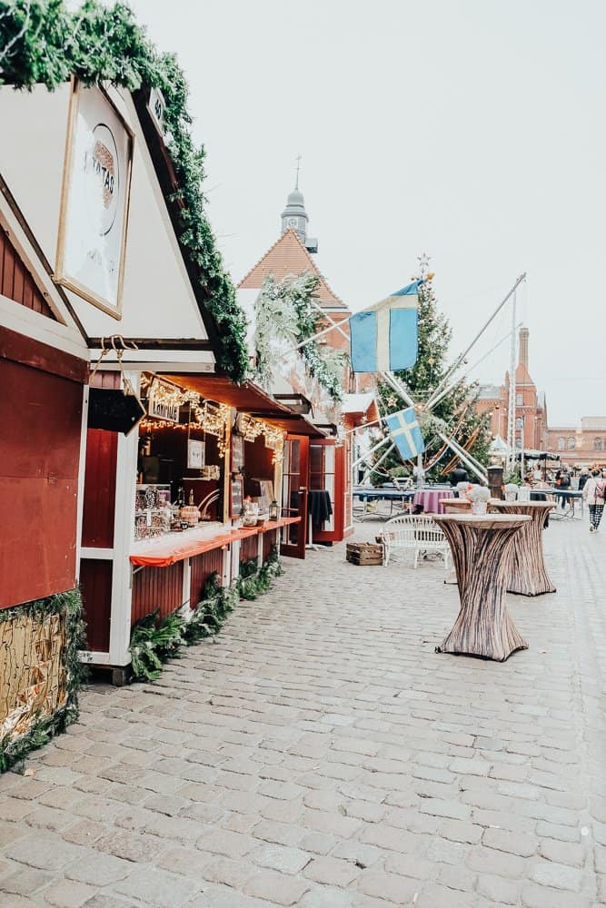 A festive market scene in Berlin, featuring wooden stalls adorned with garlands and lights, inviting passersby to explore the goods on offer amidst historic architecture.