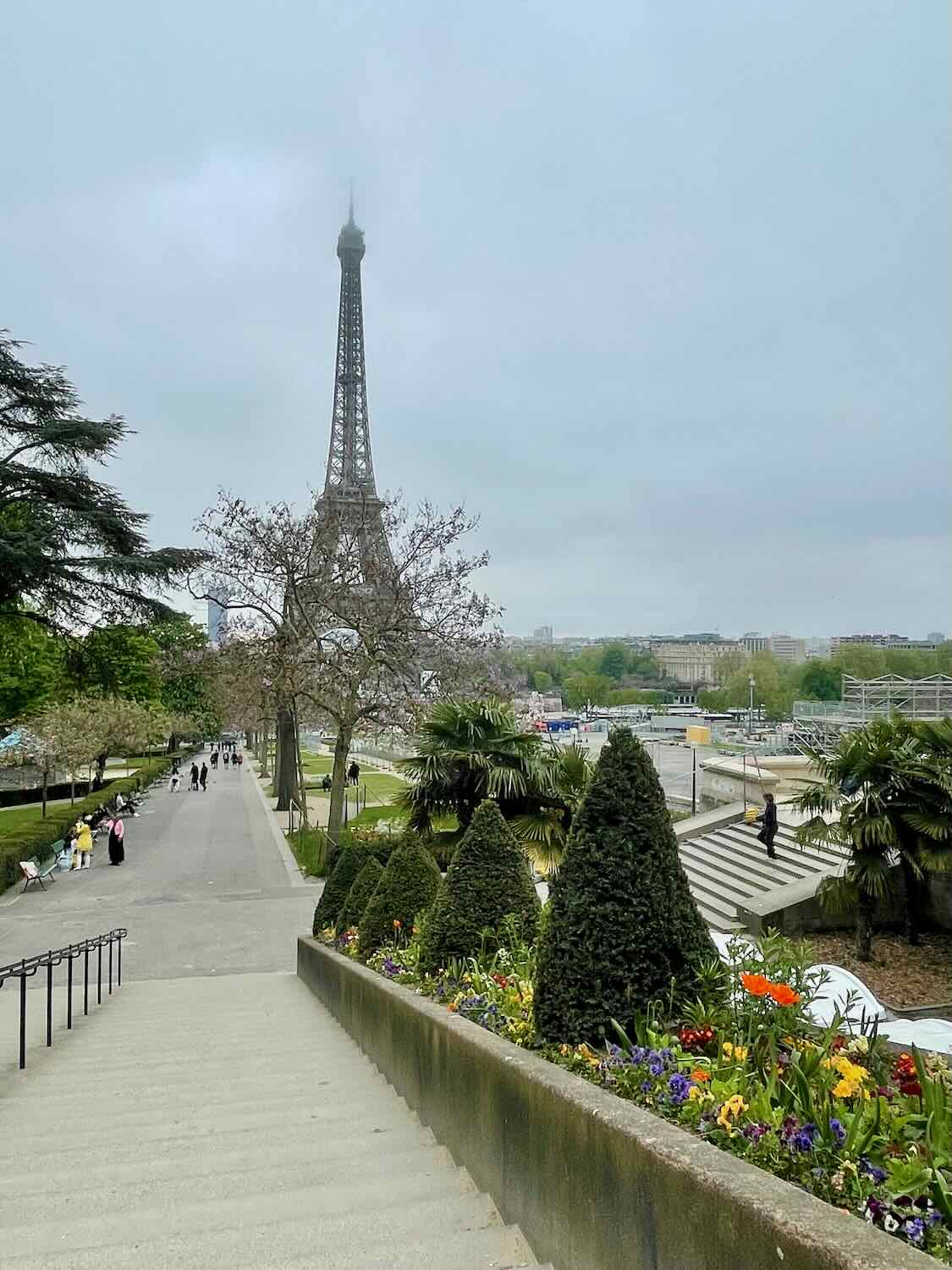 A view of the Eiffel Tower from a nearby garden path, showcasing lush greenery and colorful flowers. The overcast sky adds a dramatic backdrop to this iconic structure.