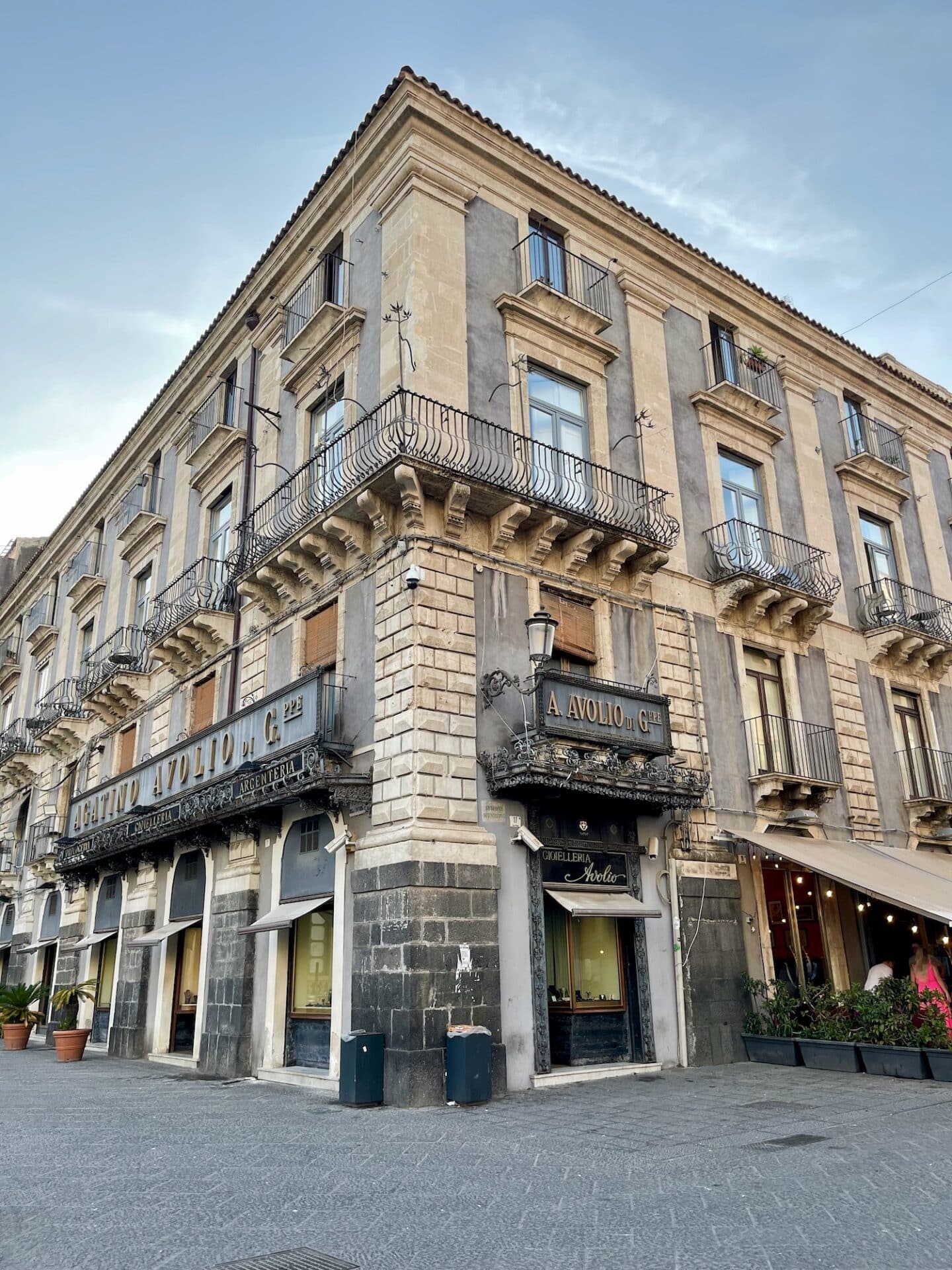 A corner building in Catania with balconies and shops at street level, under a clear sky.