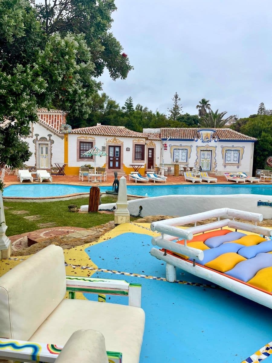 A vibrant poolside retreat in Ericeira, Portugal, featuring traditional Portuguese-style cottages adorned with intricate blue and yellow azulejo-inspired designs. The outdoor space is decorated with colorful lounge chairs, a floating daybed with multicolored cushions, and hand-painted wooden furniture. The lush greenery and blue-painted flooring contrast beautifully with the rustic terracotta tiles, creating a lively yet serene atmosphere for relaxation.