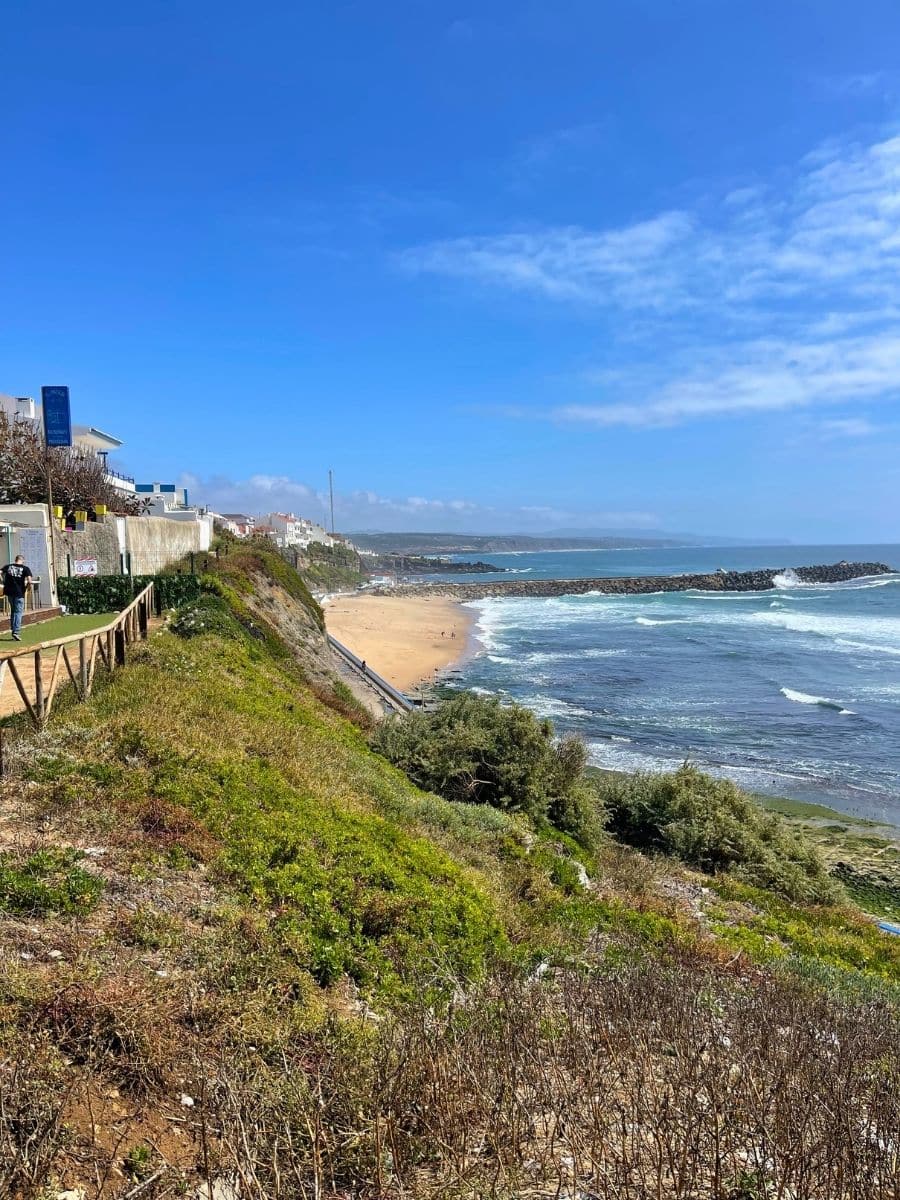 Scenic coastal view of a sandy beach and rocky shoreline in Ericeira, Portugal, with turquoise waves gently rolling in under a bright blue sky. A wooden boardwalk runs along the edge of a green cliff, where a few people are walking. Whitewashed houses perch on the hill in the distance, overlooking the Atlantic Ocean.