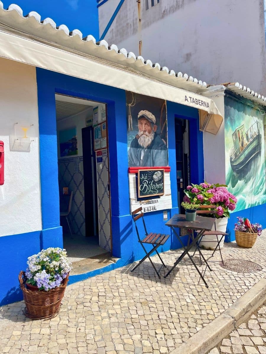 A quaint seaside tavern with bright blue and white walls, featuring a mural of an elderly fisherman smoking a pipe. Outside, a small black metal table and two chairs sit on a cobblestone street, surrounded by colorful potted flowers in baskets and planters. A sign above the door reads “A Taberna,” and a painted mural of a boat decorates the adjacent wall.