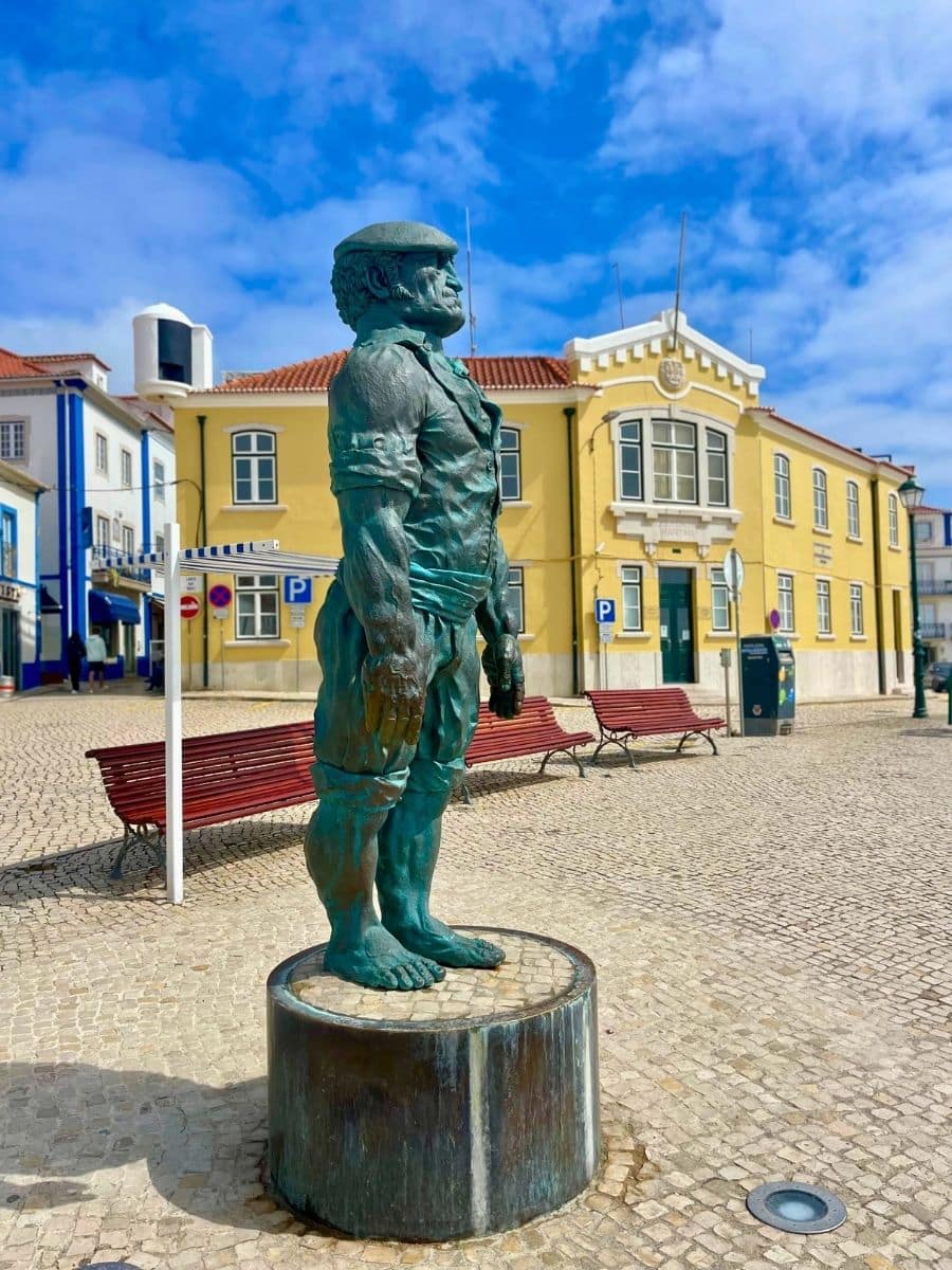 A bronze statue of a muscular fisherman in traditional clothing stands barefoot on a pedestal in a cobblestone plaza in Ericeira, Portugal. Behind the statue are red benches and colorful buildings, including a prominent yellow building with white trim and arched windows under a blue sky with scattered clouds.