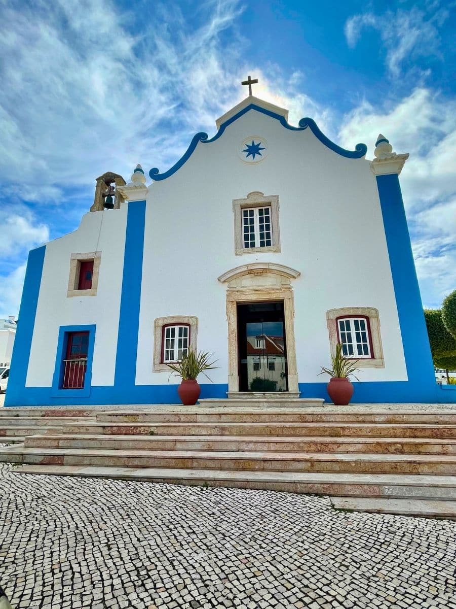 A white church with vivid blue vertical accents along its edges and a stone cross on top. The facade features a small arched window above the entrance door and a blue star-shaped symbol above it. Two potted plants sit on either side of the entrance, and the foreground is paved with a traditional Portuguese cobblestone pattern under a dramatic sky with swirling clouds.